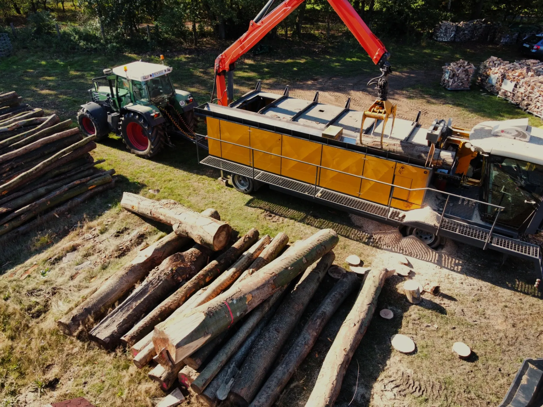 Vogelperspektive: Traktor, mobile Holzaufbereitung und Kran über Langholz auf dem Lagerplatz